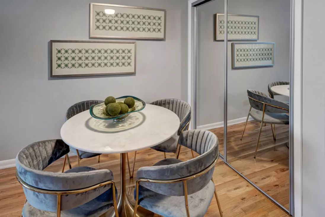 Modern dining room with a round marble table, four grey velvet chairs, green decor. Mirrored closet doors reflect the space.