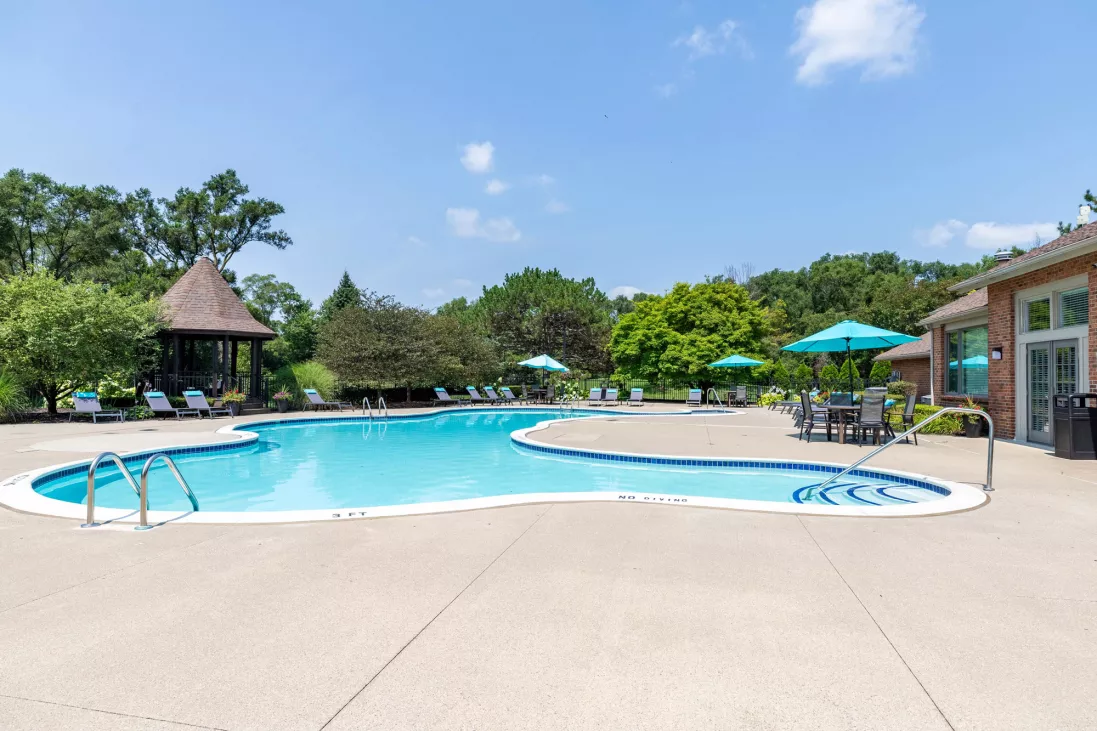 Sunny outdoor swimming pool with a concrete deck, lounge chairs, a wooden gazebo, and a nearby building, surrounded by lush trees.