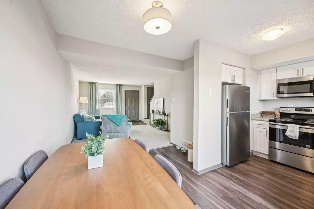 Open-concept dining area with a wooden table, leading to a kitchen with white cabinets and a living room with a blue couch.
