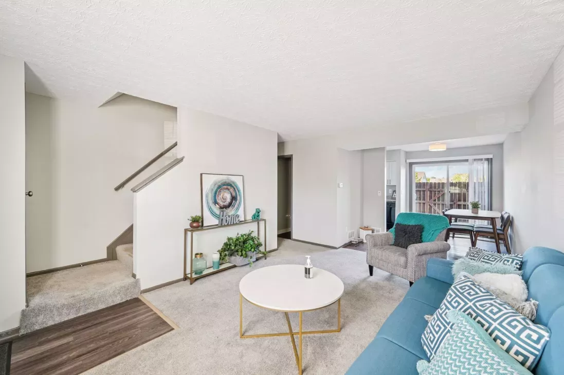 Bright, furnished living room with a light blue sofa, white coffee table, grey armchair, and a staircase leading upstairs. Dining space beyond.
