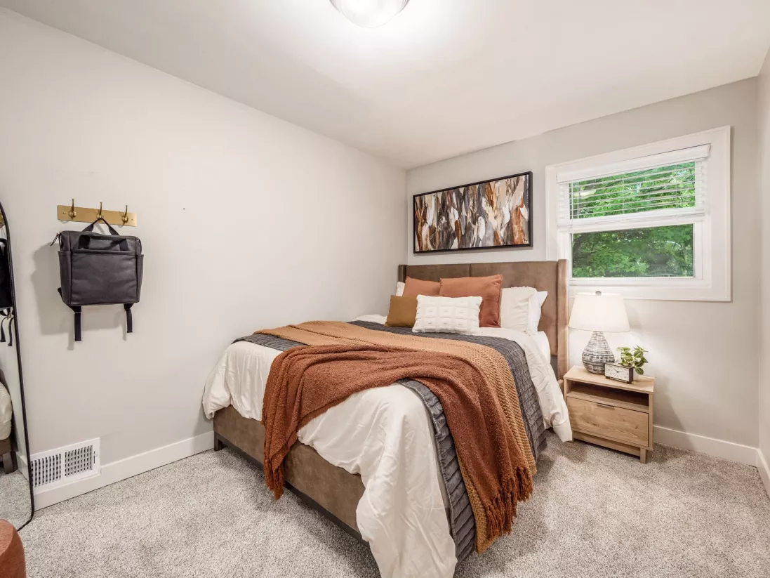 Modern bedroom with light gray walls. Bed has a white duvet, rust throw, and abstract art. Nightstand, lamp, window to trees, and a backpack.