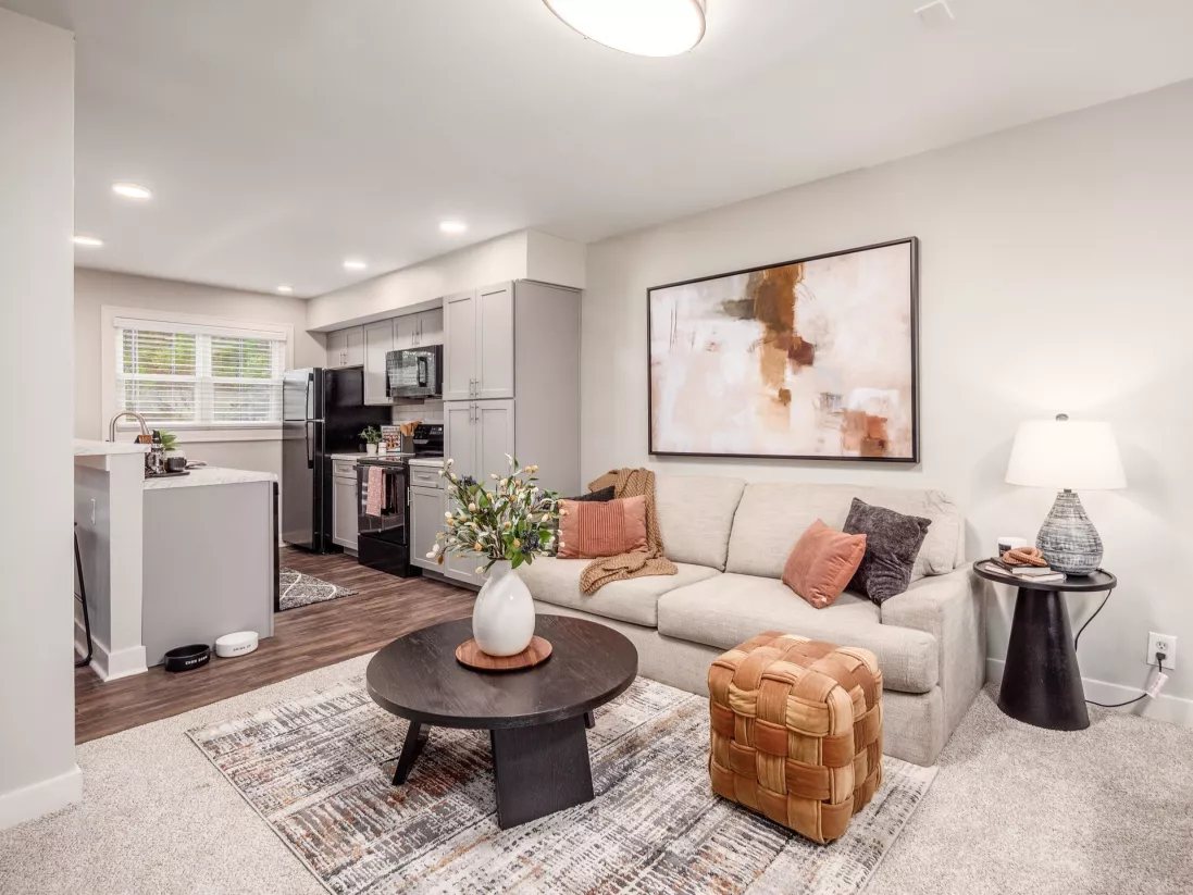 Modern living room with a beige couch, dark coffee table, abstract art, and view of a kitchen with gray cabinets.