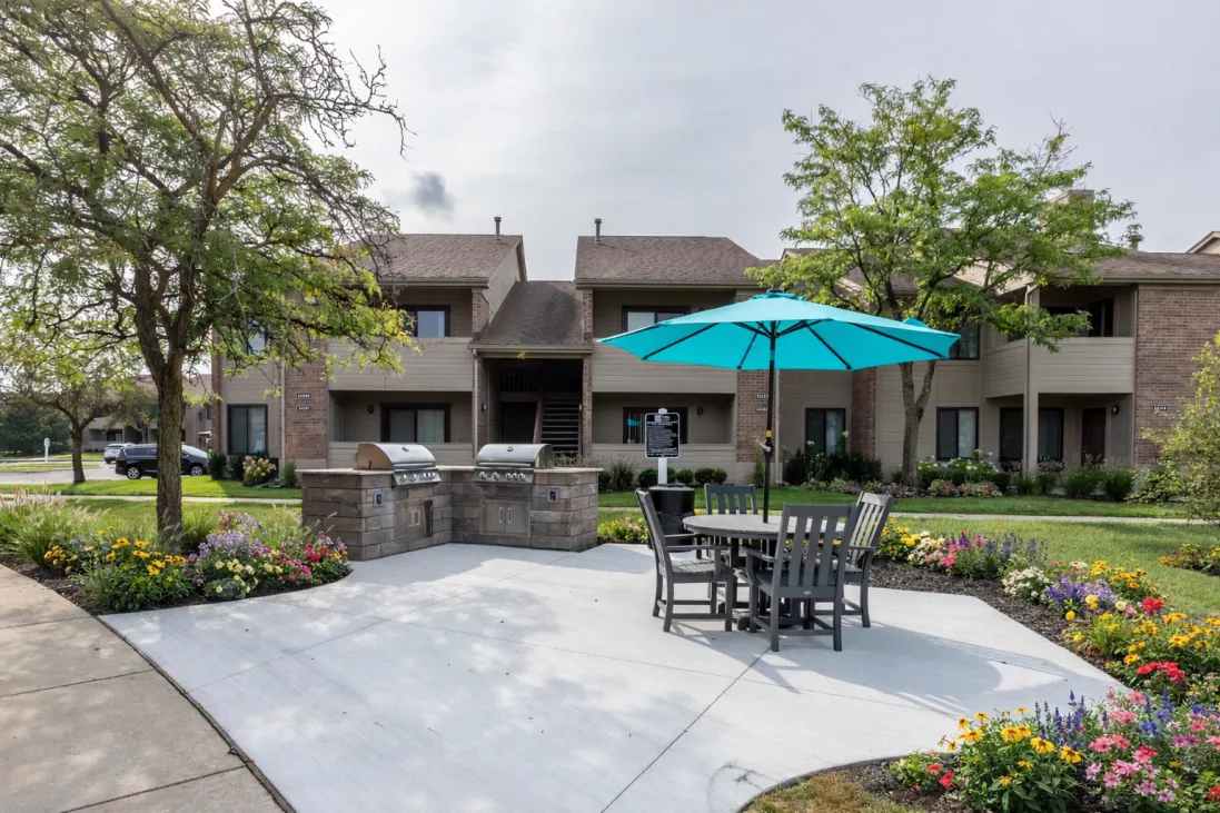 Outdoor common area with a stone grill, dining set under a turquoise umbrella, colorful flowers, and an apartment building.