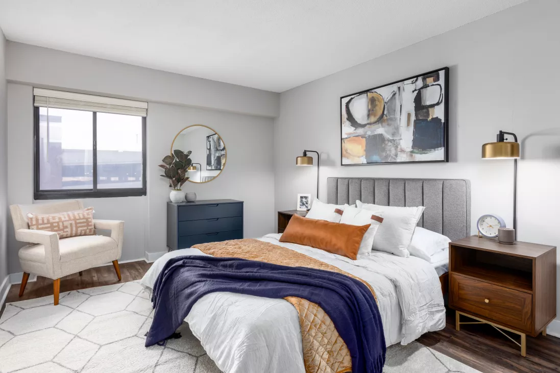 Stylish bedroom with a queen bed, grey headboard, orange and blue throws, dark wood furniture, and a white armchair.