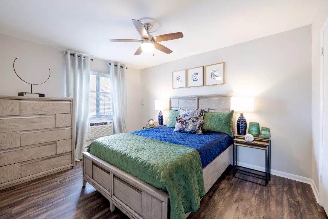 Well-lit bedroom with a bed featuring blue sheets, green throw blanket. Light wood dresser, window with blue curtains, dark wood floor.