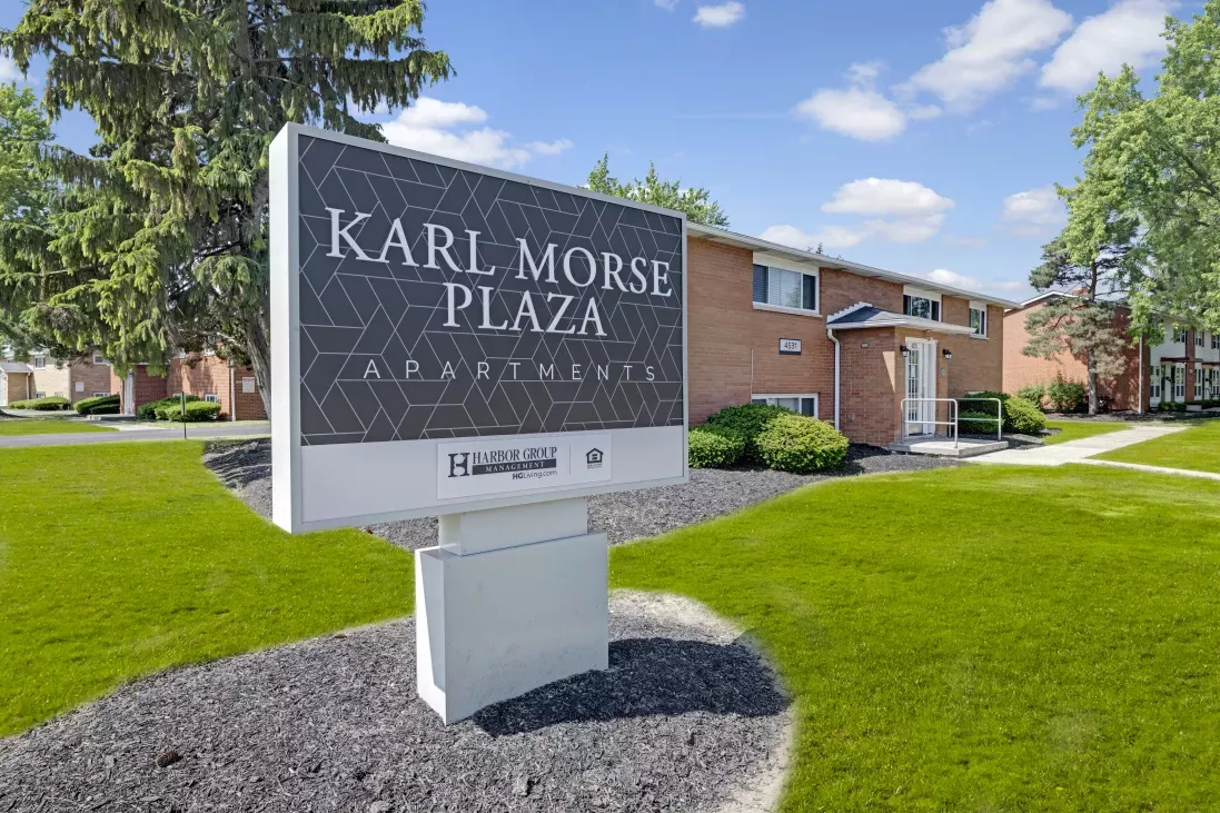 Sign for Karl Morse Plaza Apartments, a brick building with white trim, green grass, and trees under a blue sky.