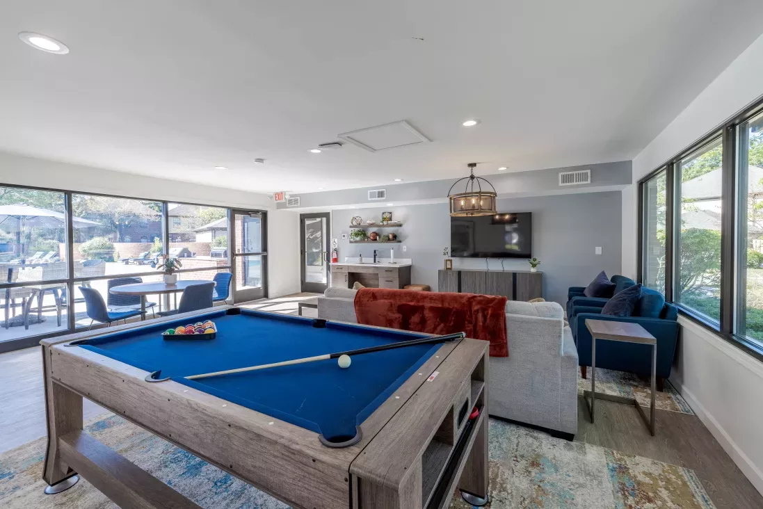 Modern common room with a blue pool table in the foreground, a lounge area with sofas and TV, and large windows revealing an outdoor patio.