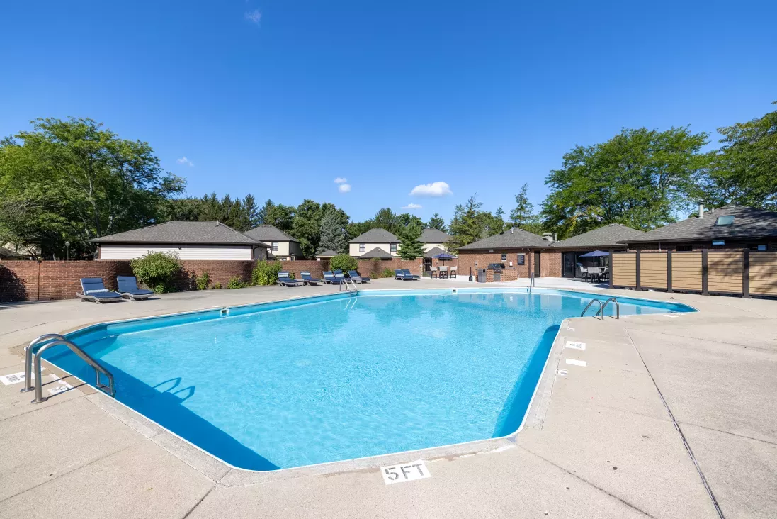 Outdoor swimming pool with clear blue water, lounge chairs on a concrete patio, and residential buildings surrounded by trees.
