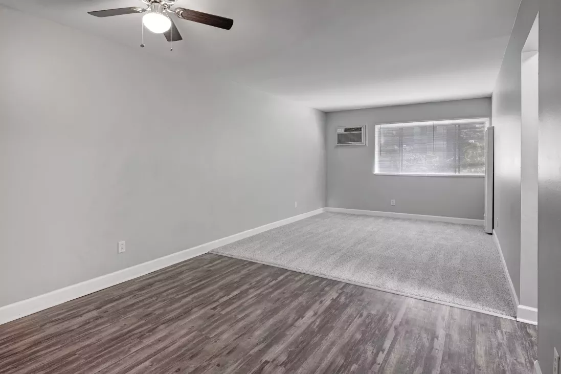 Empty room with light grey walls. Dark wood laminate floor transitions to light grey flooring, window, and ceiling fan.