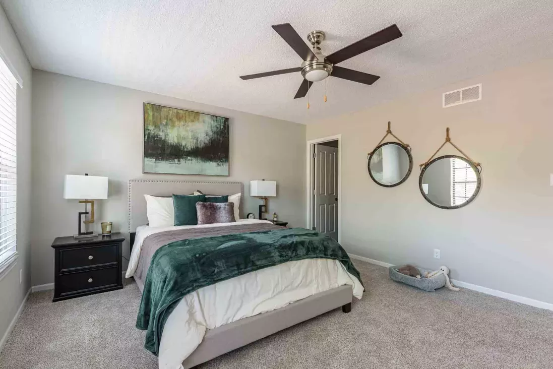 A modern bedroom featuring a bed with a dark green blanket, abstract art, two round wall mirrors, and a ceiling fan.