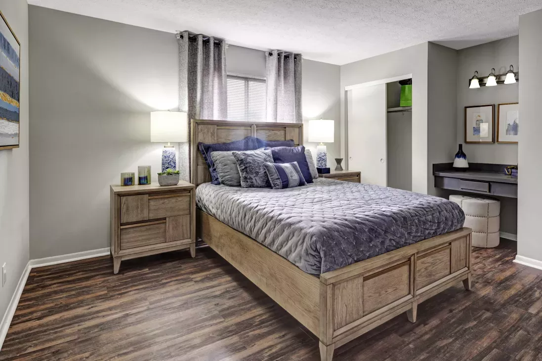 Bedroom with a wooden bed, blue and grey bedding, dark wood floor, matching nightstands with lamps, and a vanity.