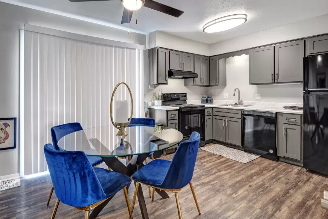 Modern kitchen and dining area with gray cabinets, dark wood floor, and a glass table with four vibrant blue velvet chairs.
