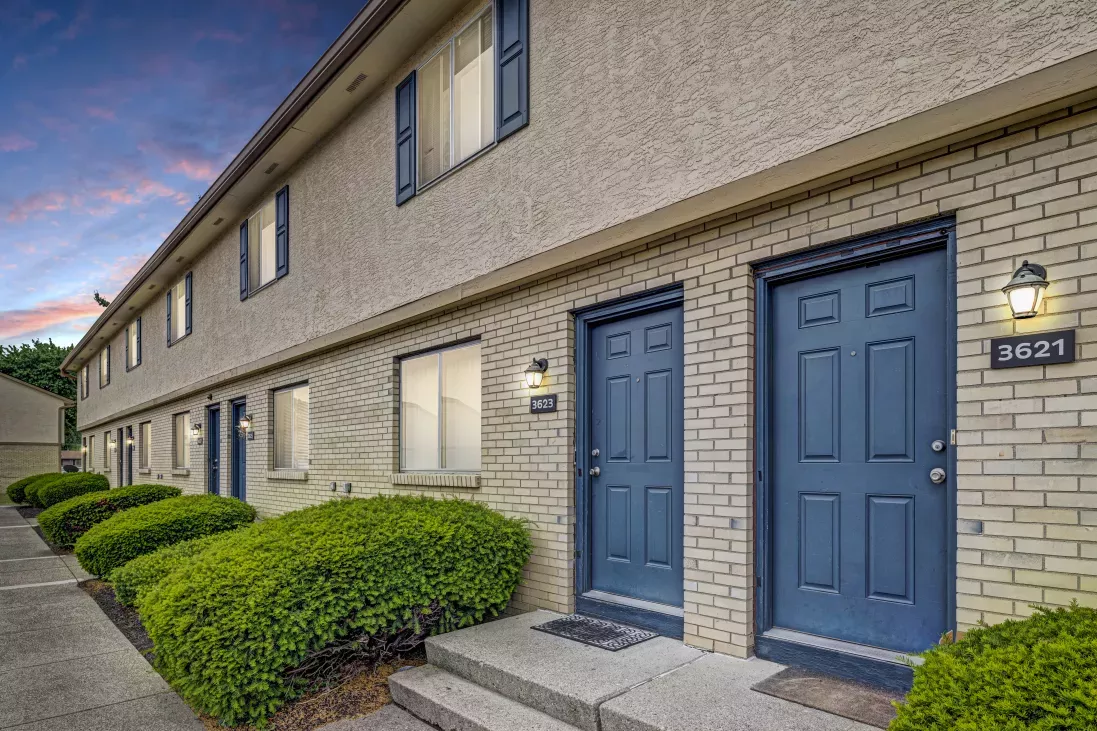 Row of apartment units with light brick, blue doors, green landscaping, and a twilight sky.