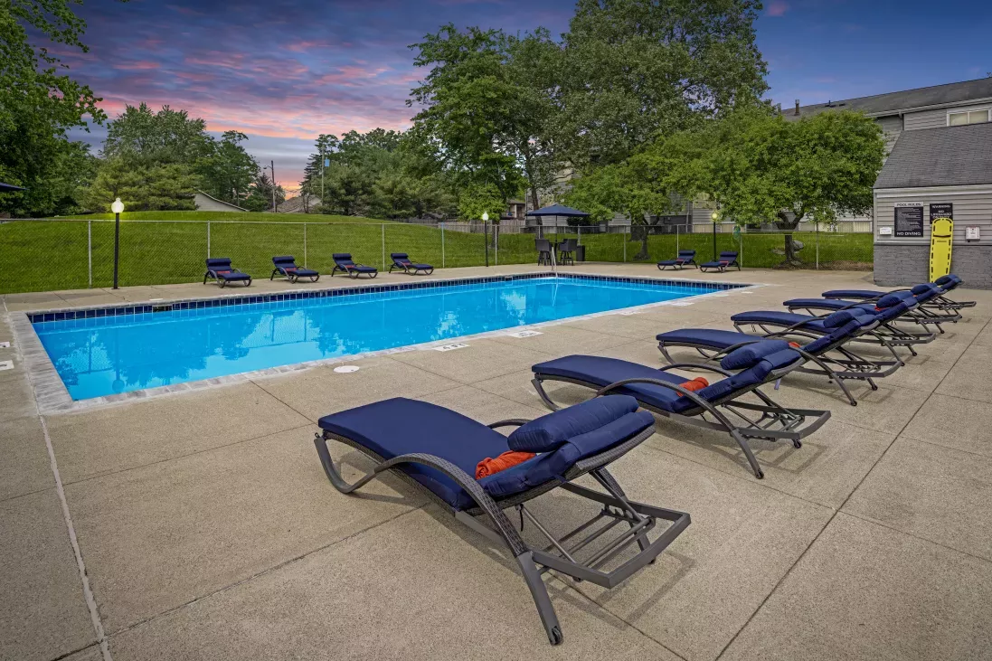 Swimming pool with blue water and lounge chairs at dusk, surrounded by trees and a building.