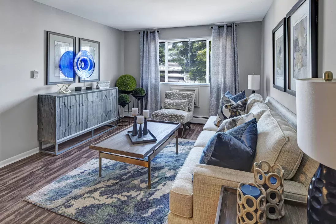 A chic living room features a white sofa, blue and white patterned rug, wooden coffee table, and credenza with blue art.