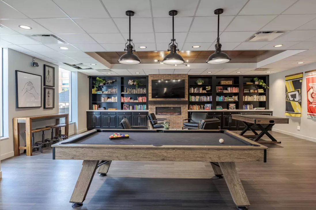 Modern lounge with a pool table, pendant lights, bookshelves, a fireplace with a TV above, and a high-top bar.