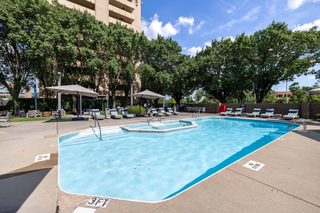 Serene outdoor swimming pool with a hot tub, lounge chairs, and umbrellas, backed by lush green trees and a building.