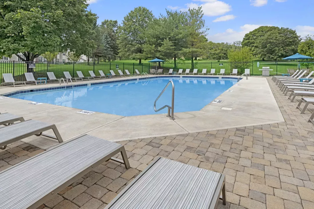 Outdoor swimming pool surrounded by gray lounge chairs on a paved patio, with trees and a blue sky.