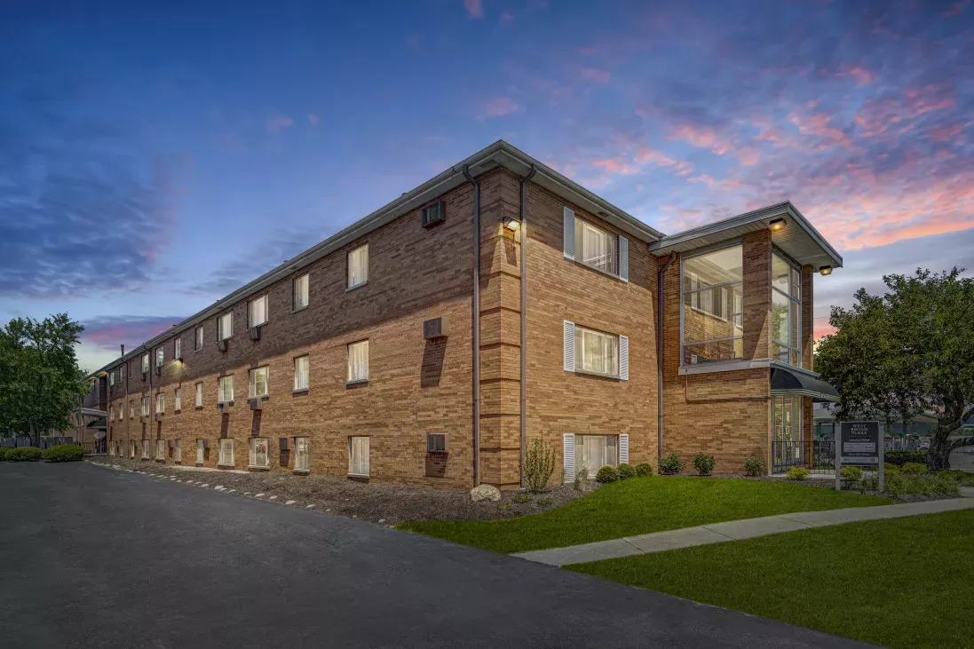 Multi-story brown brick building with a modern glass section and green lawn, viewed at dusk under a colorful sky.