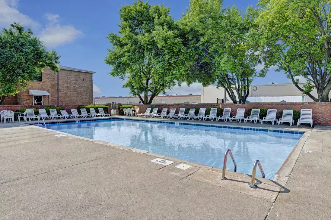 Bright outdoor swimming pool with clear blue water, many white lounge chairs on a concrete deck, and green trees.