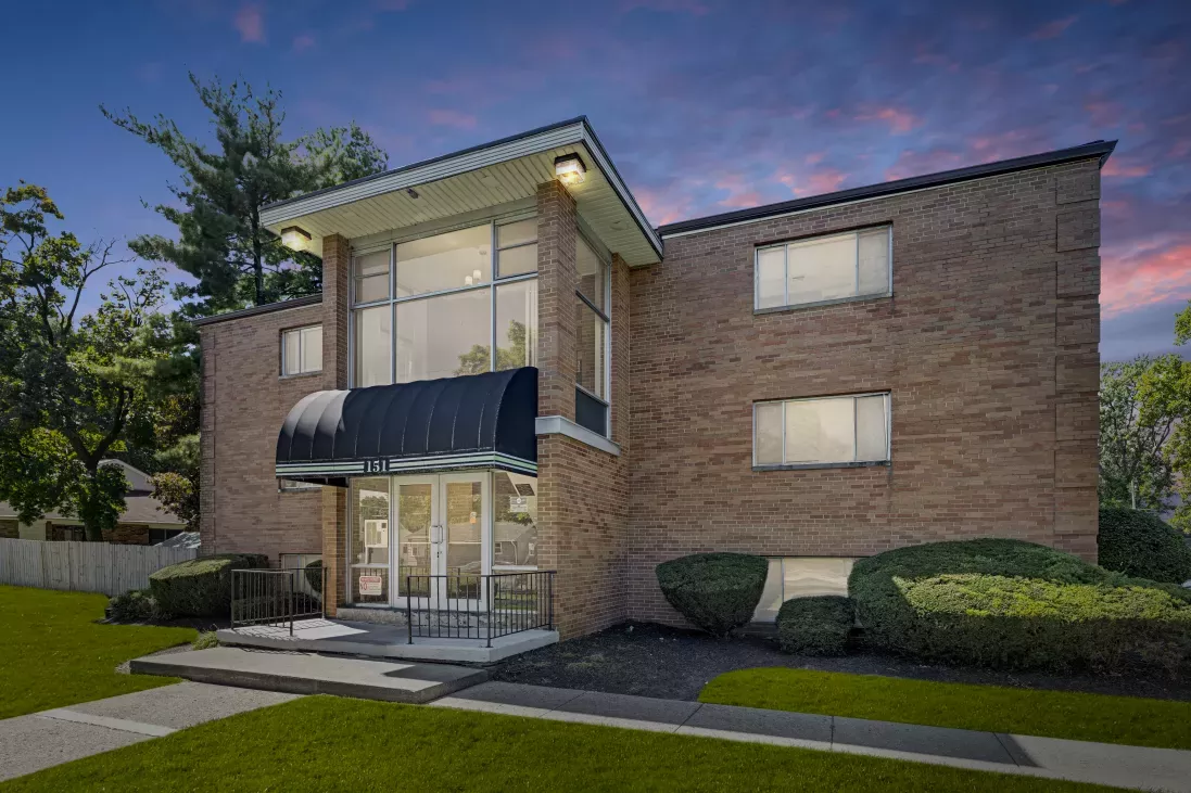 Modern two-story brick apartment building with a prominent glass entrance, black awning, green lawn, and vibrant sunset sky.