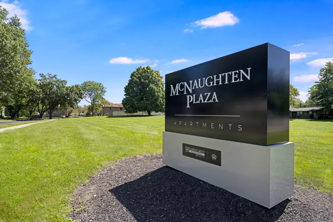 Modern black and silver sign for McNaughten Plaza Apartments on a green lawn under a bright blue sky.