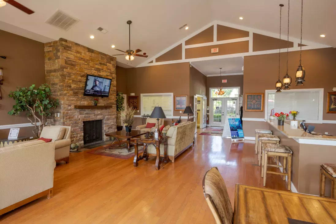 Clubhouse lounge with high ceilings, stone fireplace, TV, sofas, and a counter bar. Wood floors, warm tones.