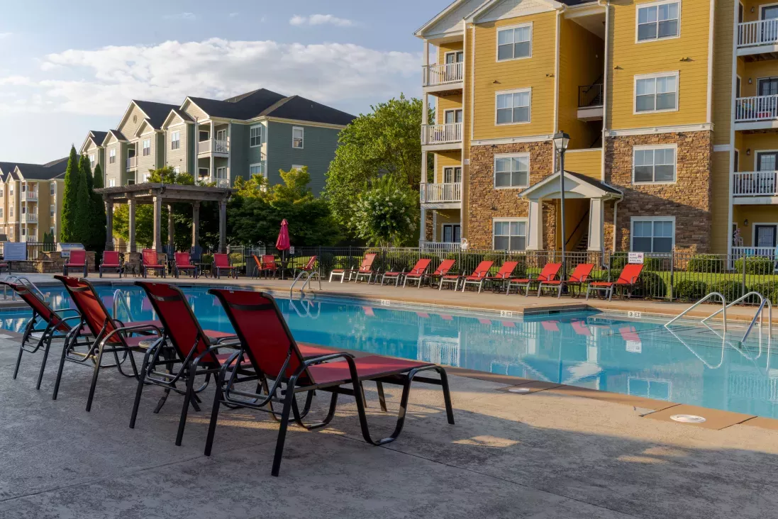 Outdoor swimming pool with red lounge chairs, bordered by yellow and green apartment buildings under a blue sky.