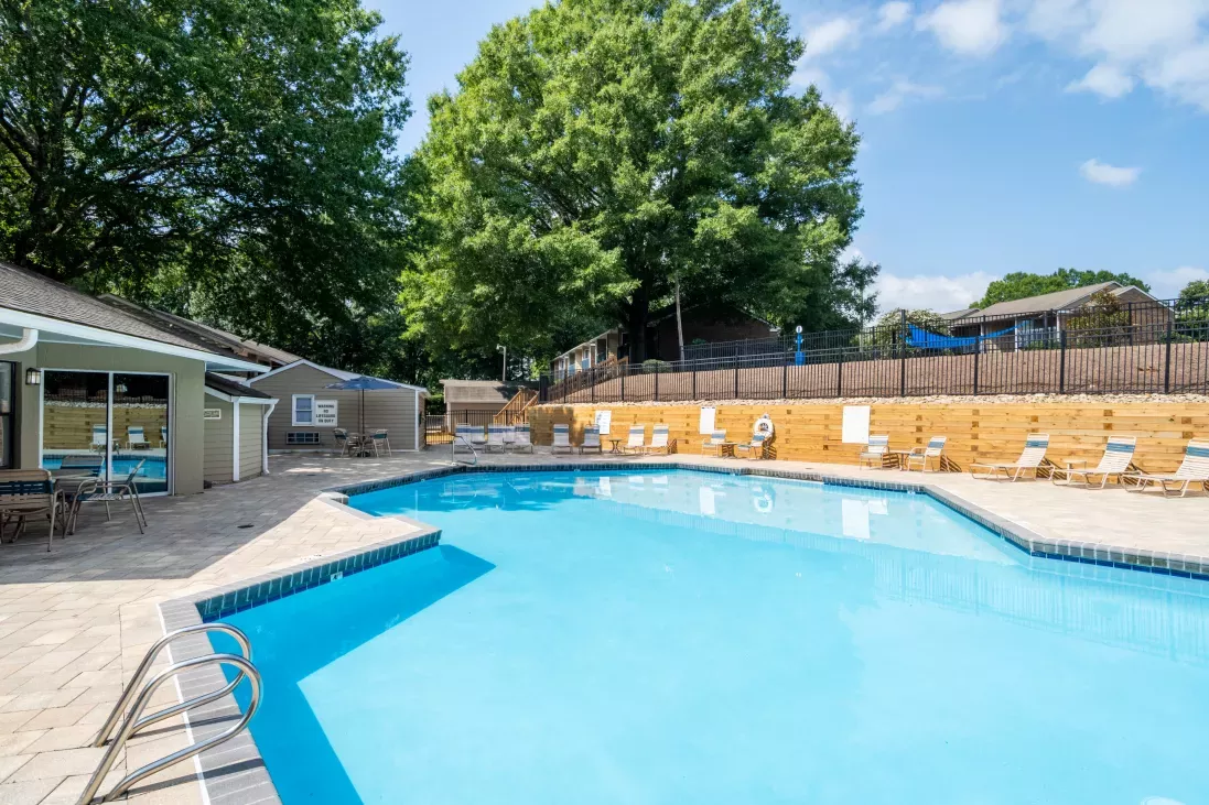 Sunny outdoor swimming pool area with a light blue pool, a tan clubhouse, and several lounge chairs.