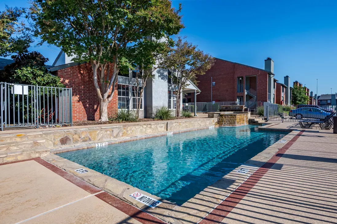 Outdoor swimming pool with a small waterfall feature in front of brick apartment buildings under a clear sky.