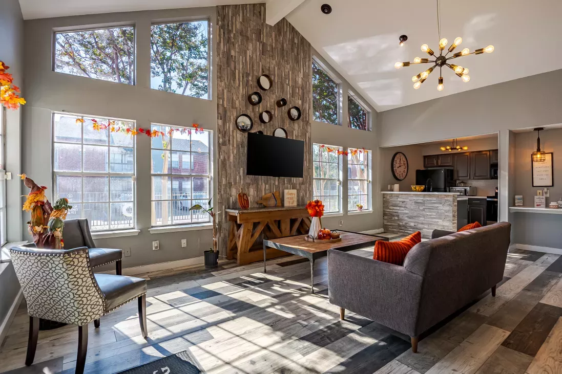 Modern lounge with high ceilings, large windows, fall decor, and gray sofas. Sunlight illuminates a patterned floor.