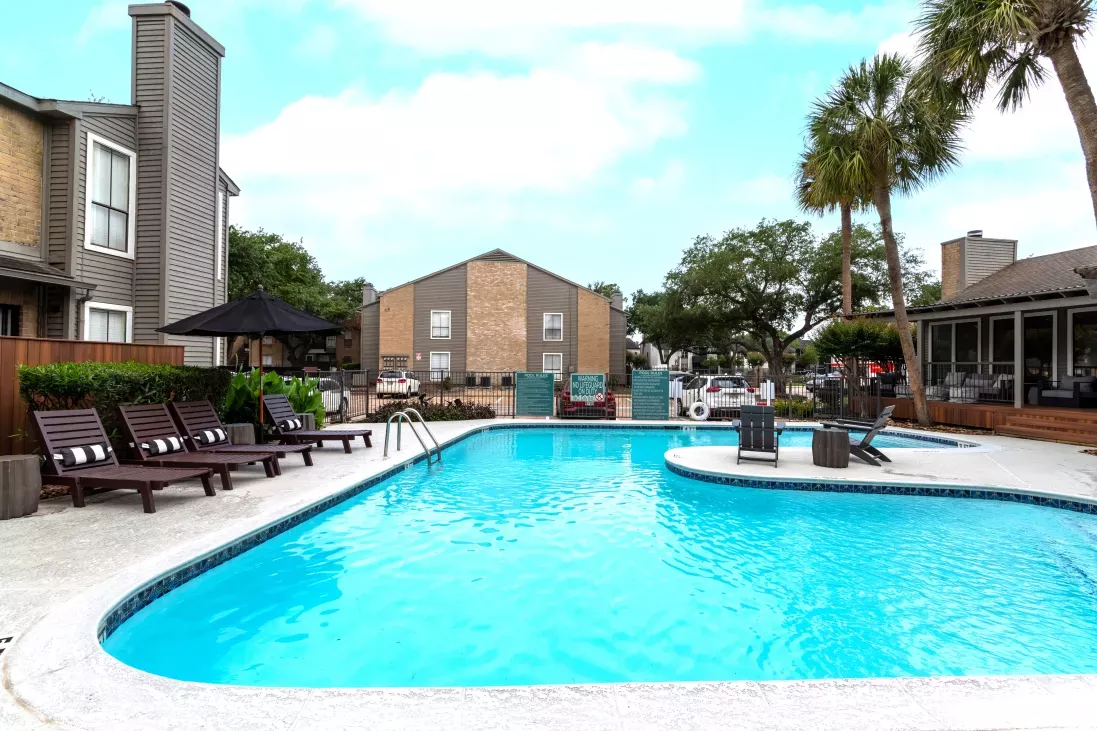 Bright blue swimming pool with lounge chairs, palm trees, and apartment buildings under a partly cloudy sky.