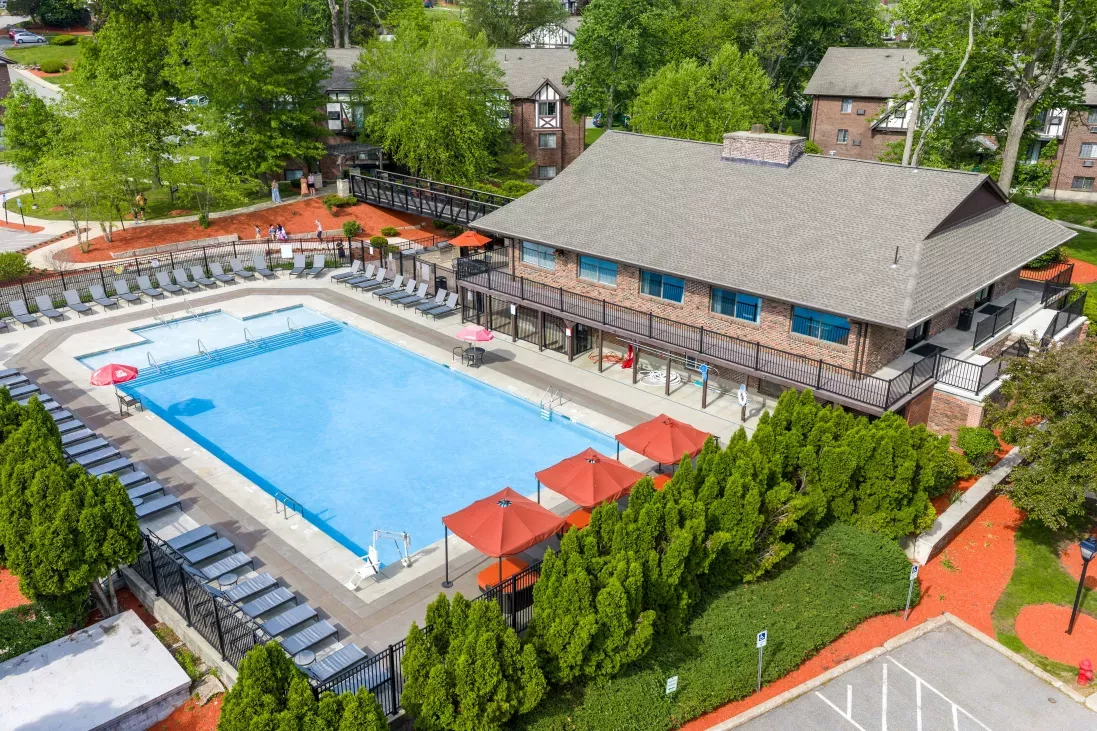 Photograph: Aerial view of an outdoor pool complex with a large blue swimming pool, lounge chairs, red umbrellas, and a clubhouse.