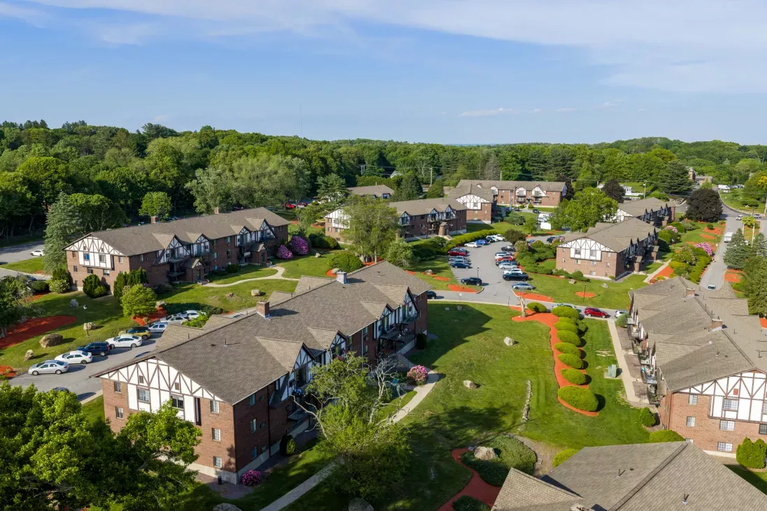 Aerial photo of an apartment complex, showing brick buildings, manicured lawns, parked cars, and surrounding trees.