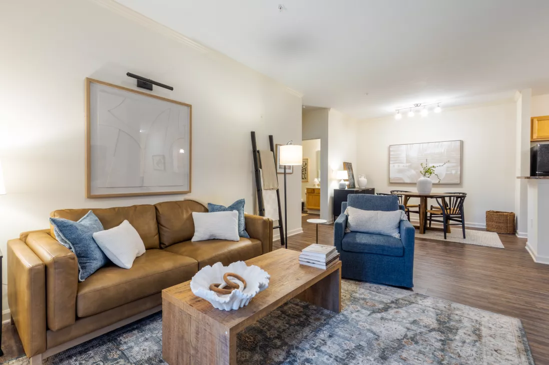 Photograph of a modern living room with a tan leather sofa, blue armchair, wooden coffee table, and an abstract rug. Dining area in background.