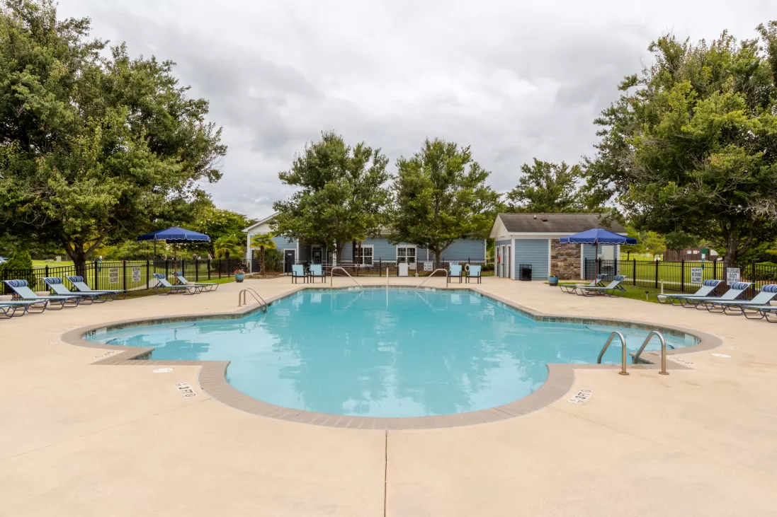 Large light blue swimming pool surrounded by lounge chairs and green trees under a cloudy sky.
