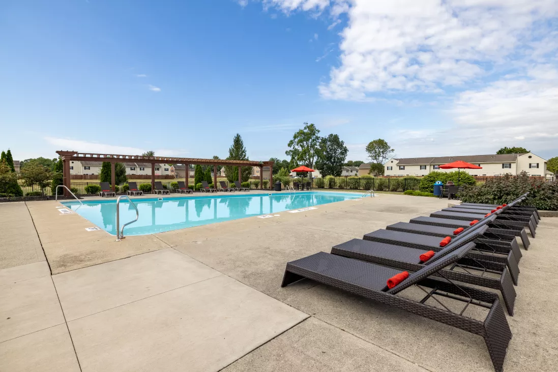 Outdoor swimming pool with many lounge chairs, surrounded by buildings and trees under a blue sky.