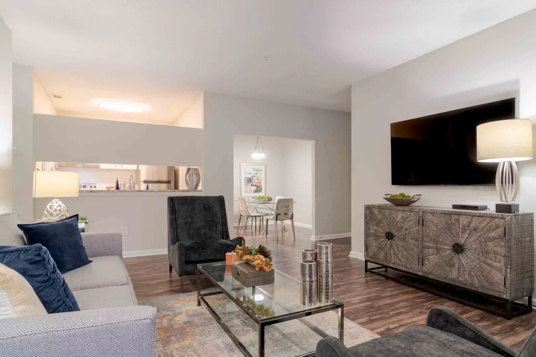 Modern living room with gray couch, dark armchairs, glass coffee table, ornate TV stand, and dining area in background.