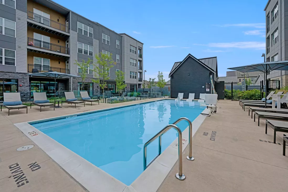 A sparkling swimming pool with poolside lounge chairs, shaded umbrellas, and contemporary apartment buildings in the background, offering a relaxing oasis for residents.