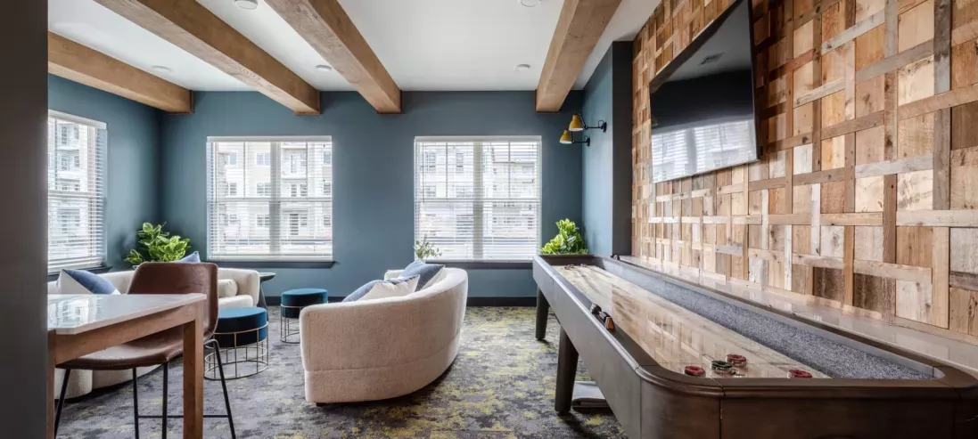 A stylish lounge area with a shuffleboard table, cozy seating, wooden accent walls, and large windows allowing ample sunlight.