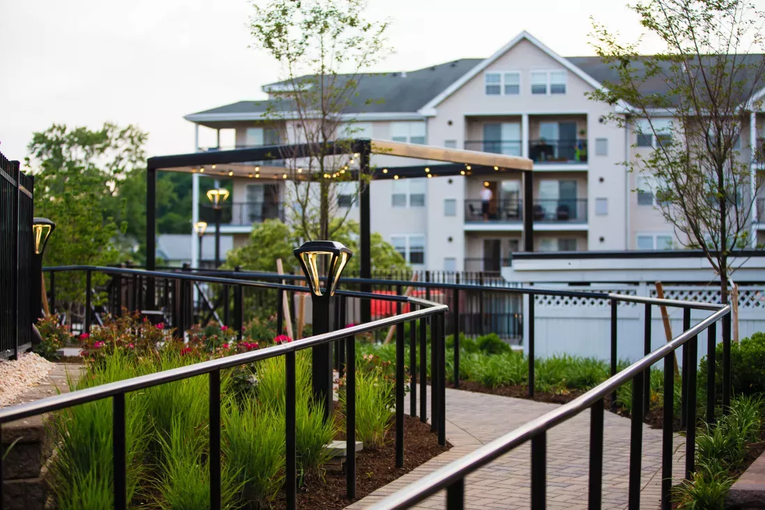 A beautifully landscaped walkway with modern railing, warm lighting, and lush greenery leading to an outdoor seating area.
