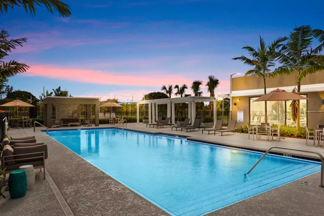 A beautifully lit outdoor swimming pool area at sunset, complete with lounge chairs, cabanas, and lush palm trees for a resort-style experience.