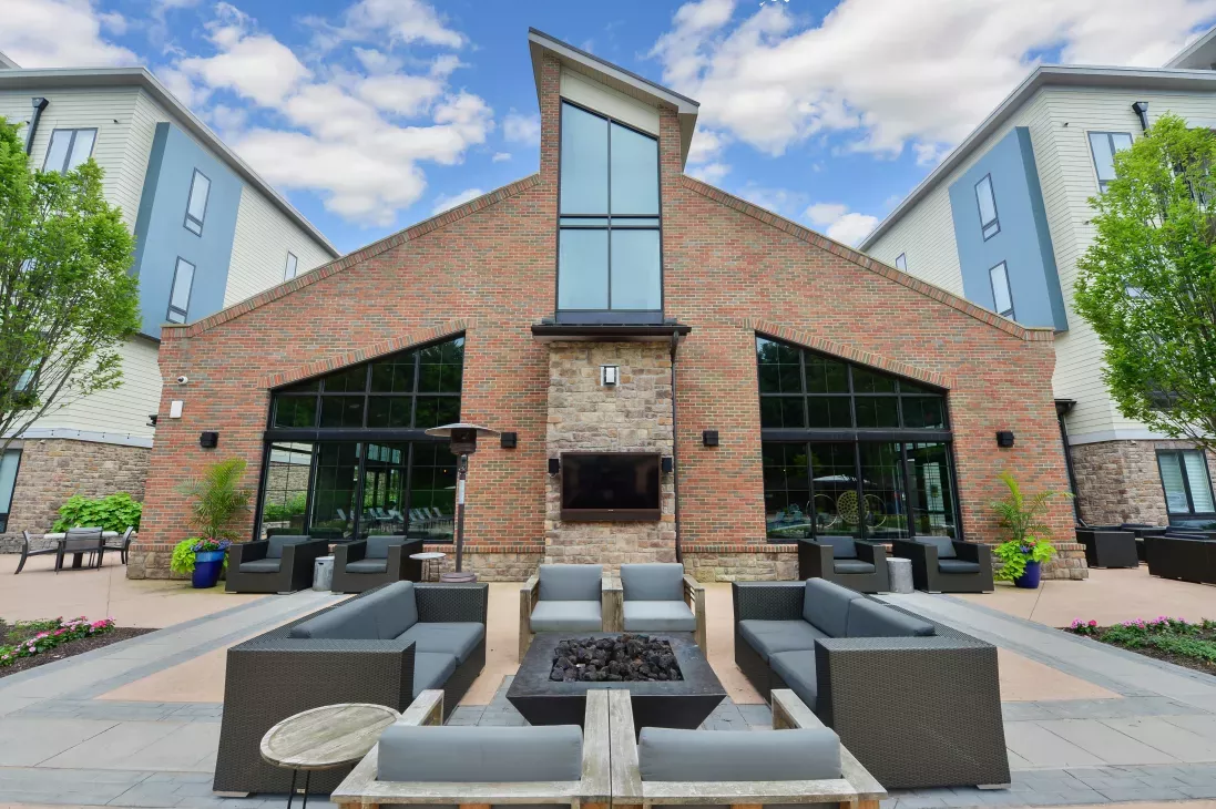 Modern outdoor lounge area featuring a brick and stone clubhouse facade, cozy fire pit, and stylish gray seating surrounded by lush greenery.