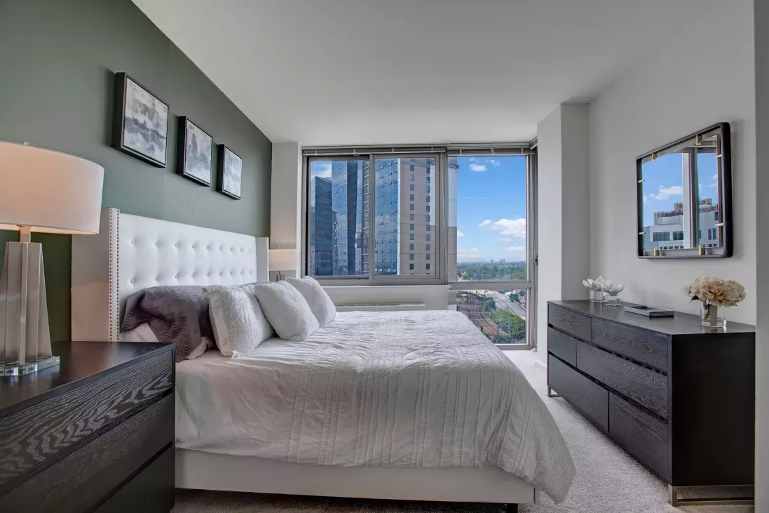 Luxurious bedroom at Skyline New Rochelle with a tufted headboard, elegant furnishings, and large windows showcasing scenic urban vistas.