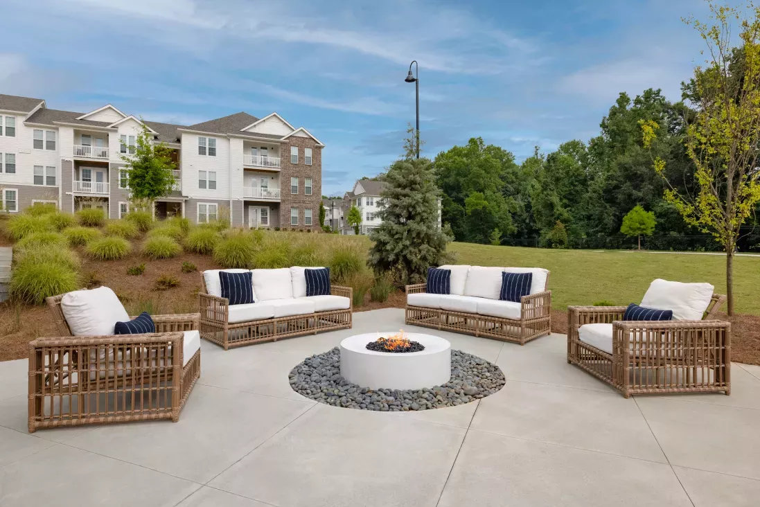 A stylish outdoor fire pit lounge featuring plush wicker seating with white cushions and navy accent pillows, surrounded by lush landscaping and modern apartment buildings in the background.