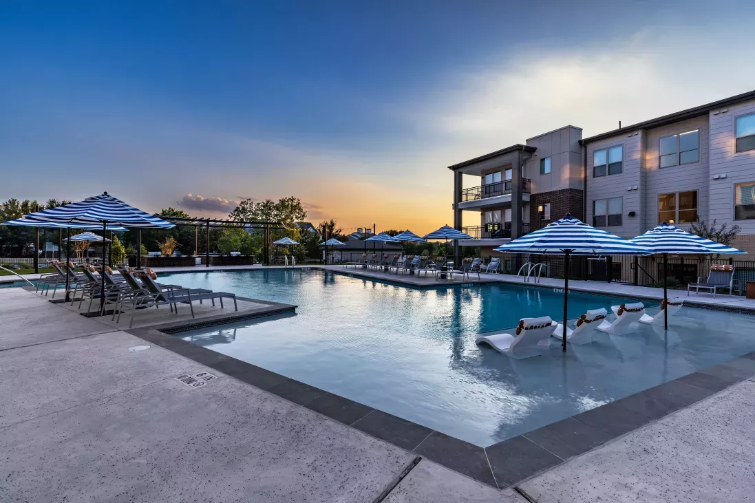 A breathtaking view of The Warner Apartments' pool at sunset, with in-water loungers, striped umbrellas, and elegant lighting creating a serene ambiance.