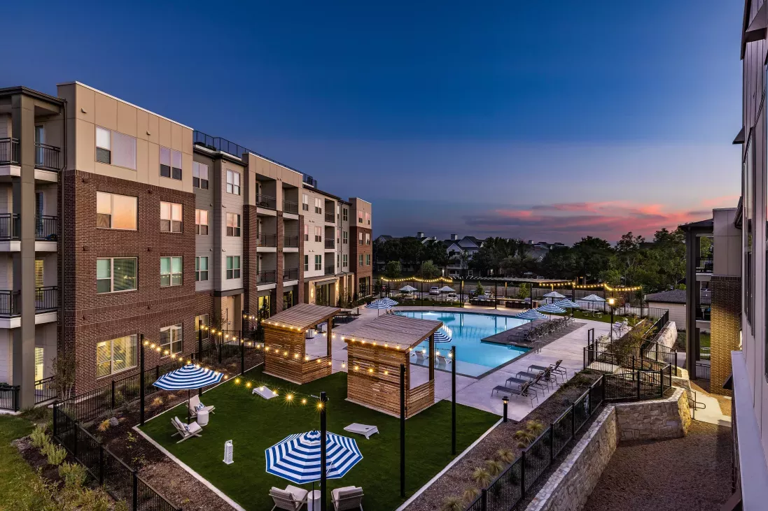 An aerial view of The Warner Apartments' pool and courtyard at dusk, featuring glowing string lights, shaded lounge areas, and a well-maintained green space.