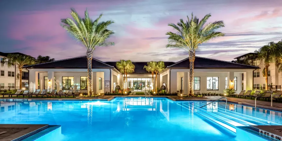 A luxurious resort-style pool illuminated at dusk, surrounded by palm trees and lounge chairs.