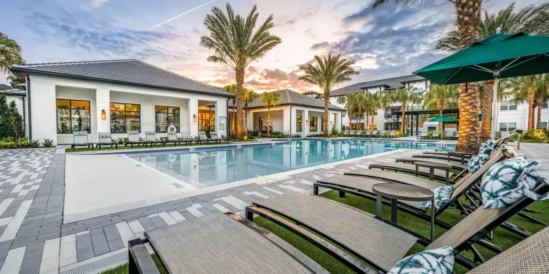 A serene poolside area with cushioned lounge chairs, umbrellas, and tropical landscaping at sunset.