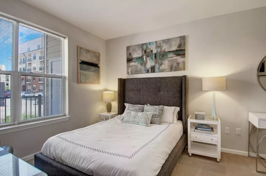 Cozy bedroom with gray upholstered bed, neutral decor, and two windows providing exterior views.
